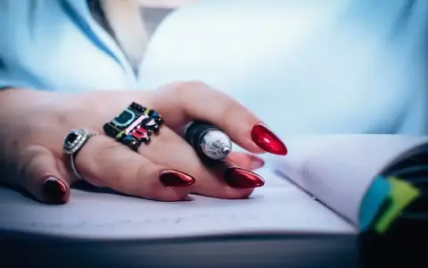 Dłoń z czerwonymi, lśniącymi paznokciami i pierścionkami trzyma długopis nad notatnikiem. To dowód, że witaminy na paznokcie działają!