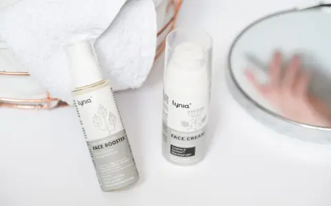 Białe opakowania kosmetyków Lynia: Face Booster i Face Cream, z widocznym składnikiem papaina. Obok ręczniki i lustro.