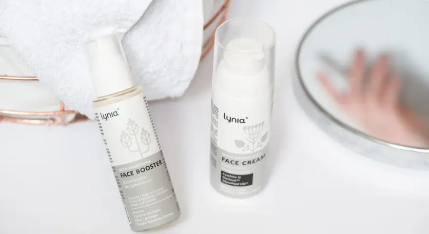 Białe opakowania kosmetyków Lynia: Face Booster i Face Cream, z widocznym składnikiem papaina. Obok ręczniki i lustro.